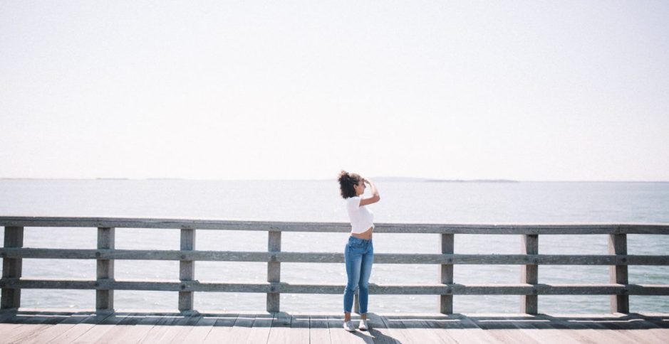 A woman in a white top and blue jeans stands on a wooden deck overlooking a calm ocean on a bright day.