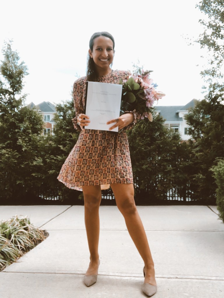 A smiling woman stands outdoors holding a document and a bouquet of flowers, wearing a patterned dress and beige heels.