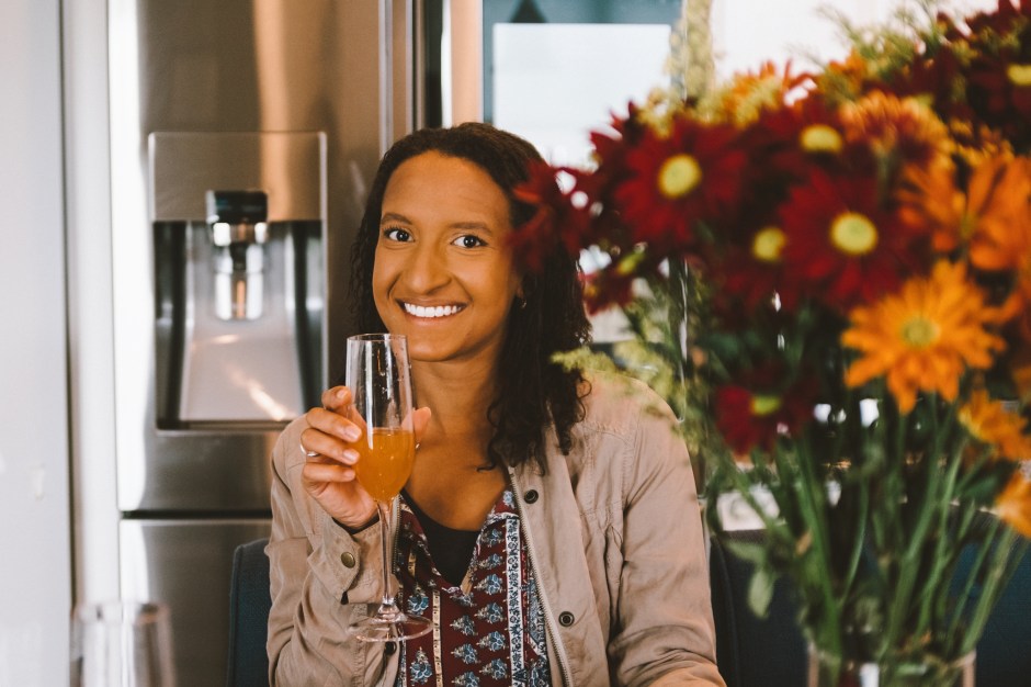 A smiling woman holding a glass of orange beverage, sitting at a table decorated with colorful flowers in the foreground, with a kitchen in the background.