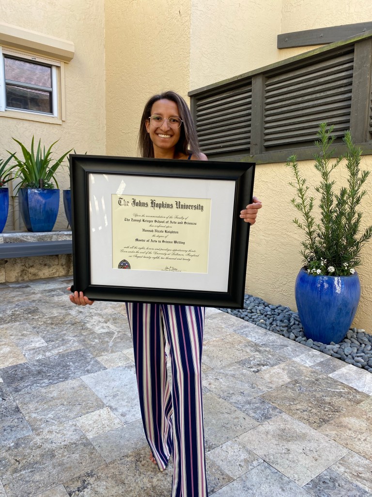 A young woman smiling and holding a framed diploma from Johns Hopkins University, standing outdoors in a patio area with potted plants.