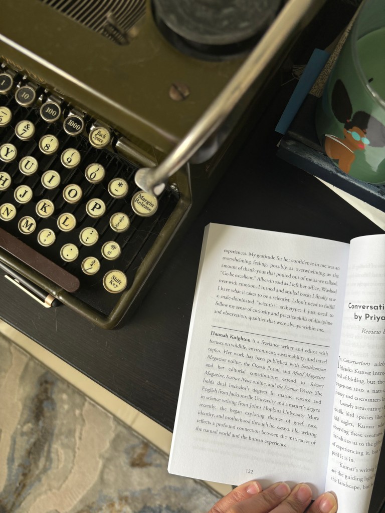 An overhead view of a vintage typewriter next to an open book, with a person's hand visible holding the book. A decorative mug is partially seen in the background.