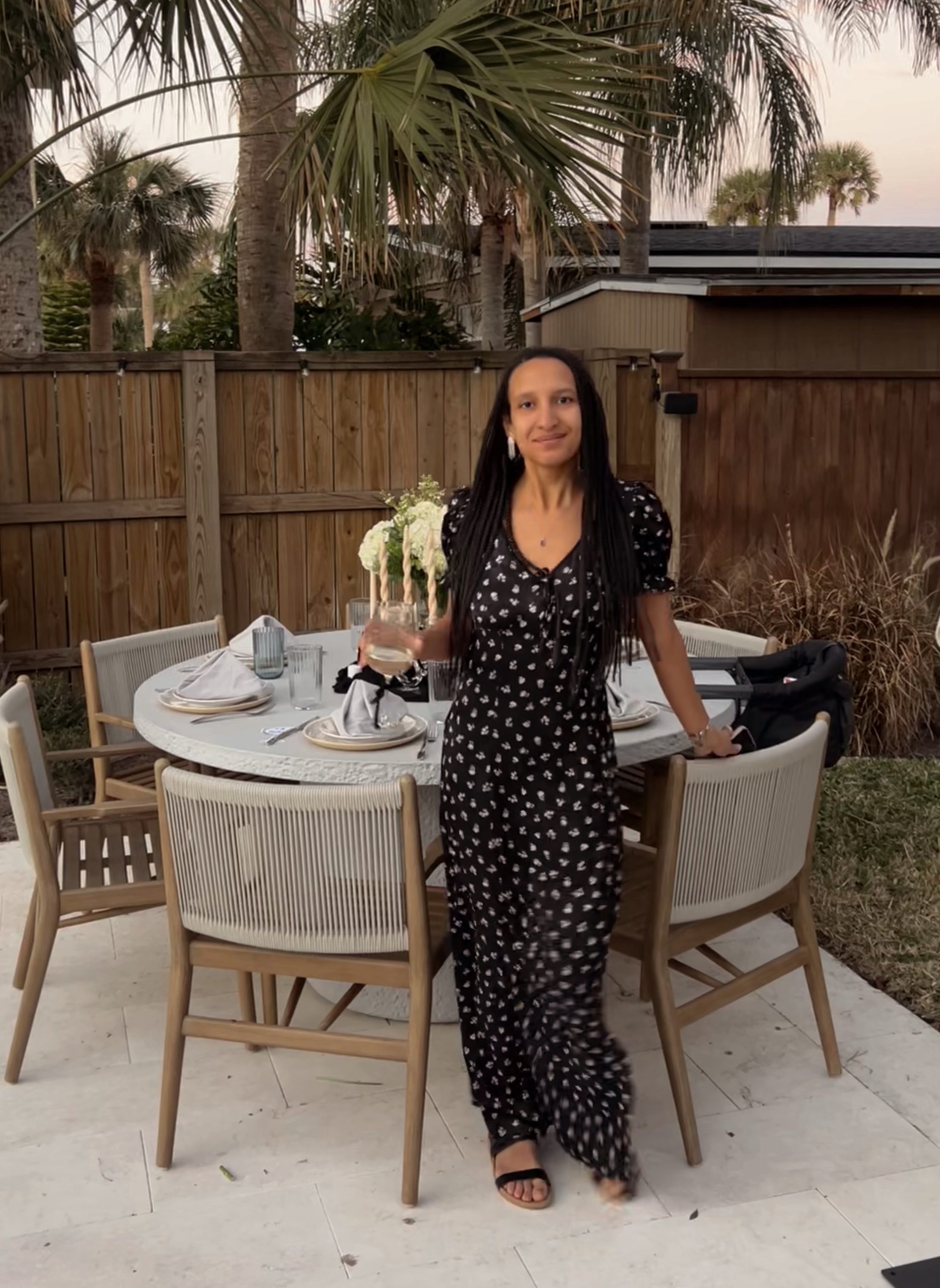 A woman standing next to a dining table in an outdoor setting, wearing a black floral dress and holding a drink, with palm trees and a wooden fence in the background.